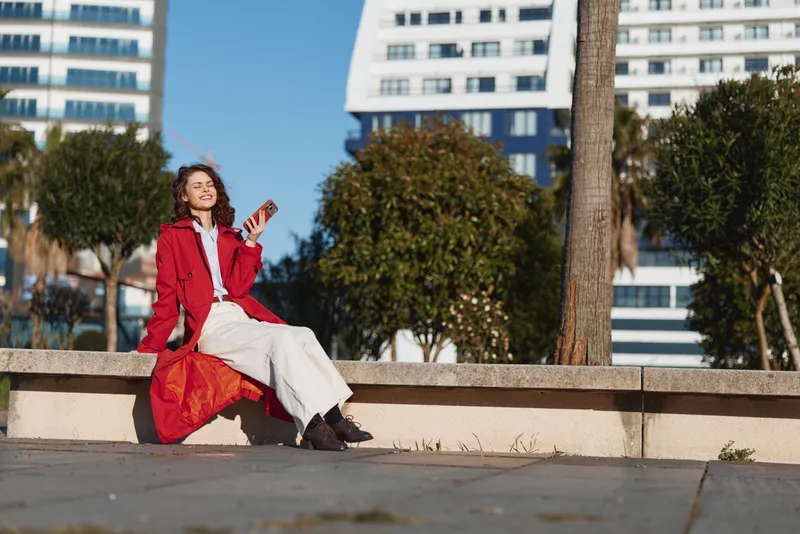 Young woman in a red coat sits on a stone bench outdoors enjoying sunlight while holding a smartphone and smiling with closed eyes.