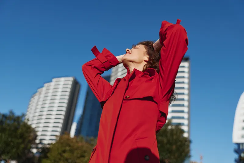 A person in a bright red coat tilts back with arms raised, enjoying a sunny day in an urban park as tall buildings loom in the background.