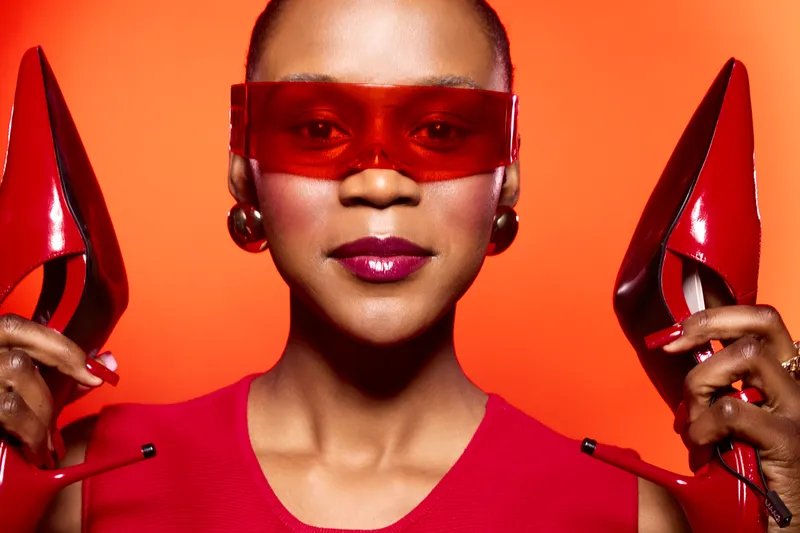 Confident black woman with dark skin wearing red sunglasses and red dress holds red high heels near her face. Bright orange background creates vibrant fashion and style concept.