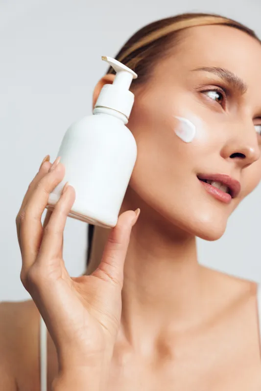 Young woman with blonde hair applies moisturizing cream on her cheek while holding a white lotion bottle against a light background, expressing a calm and fresh skincare moment.