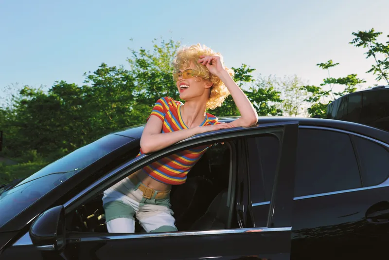 woman lifestyle scene with retro film color palette, smiling woman leaning through car window, outdoor sunlight, striped shirt, candid portrait of carefree joy and casual fashion