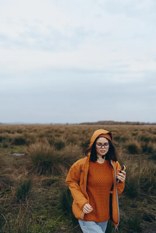Woman lifestyle outdoors field with tall grass, orange jacket and beanie, standing on a path and holding hair, windy evening light, calm nature travel mood