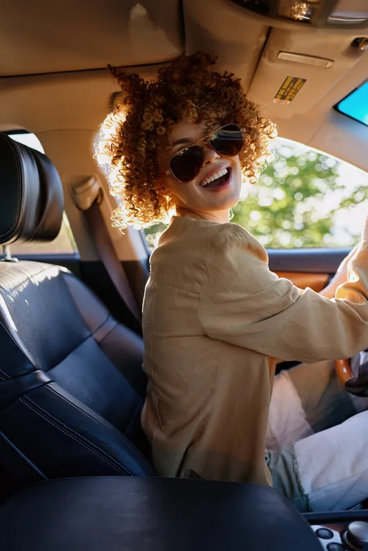 A woman with curly hair wearing sunglasses sits in a car, smiling at the camera, capturing a carefree, upbeat lifestyle moment with retro film color and warm natural light.