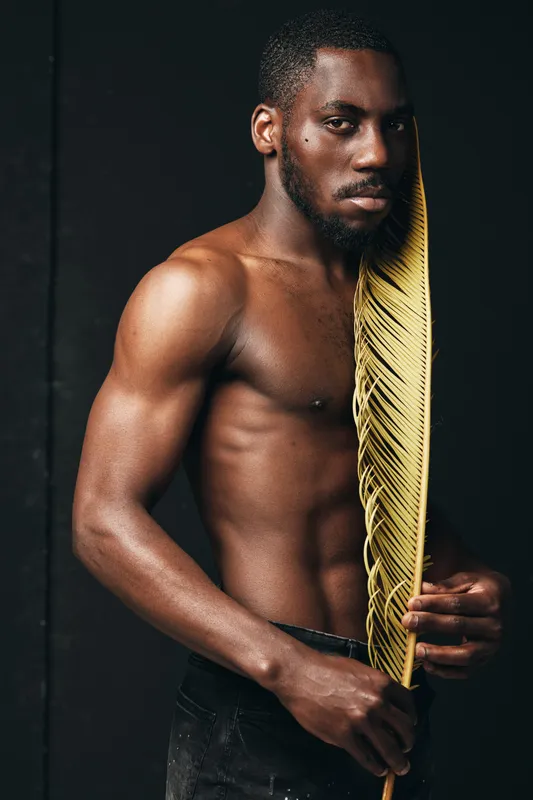 African man with muscular body and serious expression holding large yellow leaf in creative conceptual visual style. Dark skin, bare torso, artistic portrait with black background and dramatic