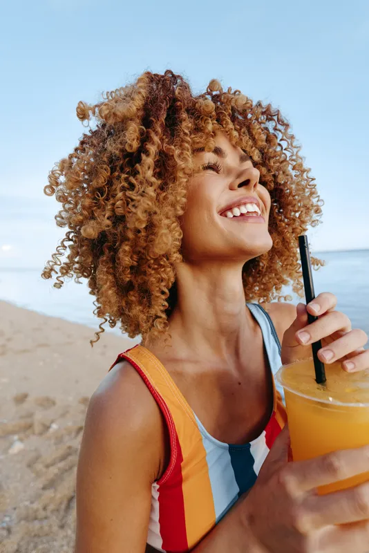 Young woman with curly hair on a sunny beach smiles while drinking orange juice from a bottle, wearing a striped swimsuit as the sea and sand glow behind her in a vibrant summer moment.