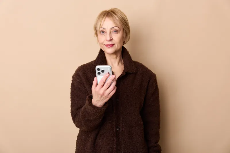 Senior woman portrait indoors holding smartphone, looking at camera with calm friendly expression, soft studio light on beige background, digital communication and technology theme
