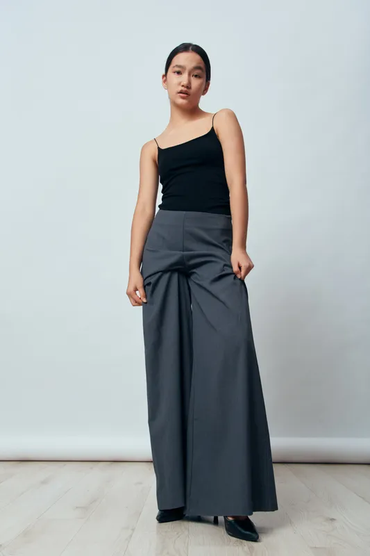 Young woman in black tank top and wide leg gray pants standing on wooden floor with neutral expression. Fashionable casual outfit with simple background for style and clothing inspiration.