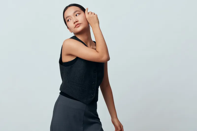 Young woman posing with hand near face, looking away and wearing sleeveless black top and gray skirt against light background in studio. Female model with natural makeup and confident expression.