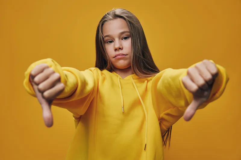 Girl in yellow hoodie showing thumbs down with both hands, looking serious and dissatisfied on solid orange background with casual long hair style.