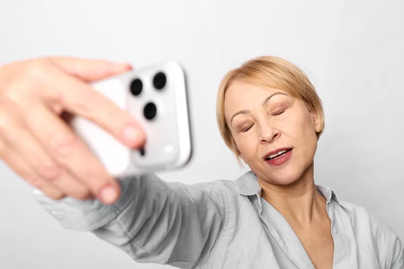 Woman holding a smartphone takes a selfie during a video call, supporting communication and technology, with a soft portrait mood of beauty, wellness, and skincare, calm studio background.