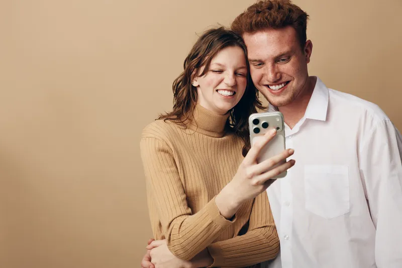 Smiling young couple takes selfie with smartphone on beige background. Casual dressed man and woman enjoy moment together, showing happiness and connection in modern lifestyle concept.