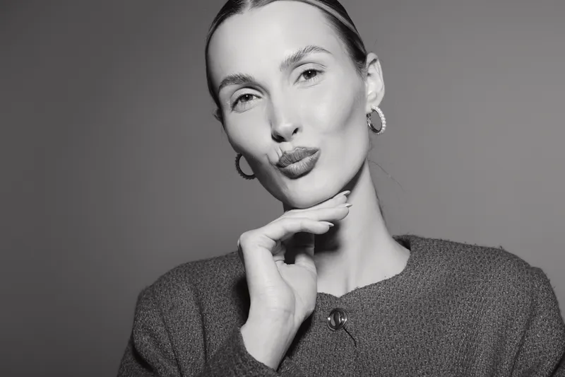 Young white woman with light skin and slicked back hair blows a kiss with closed eyes. Studio portrait in black and white with soft lighting and casual sweater.