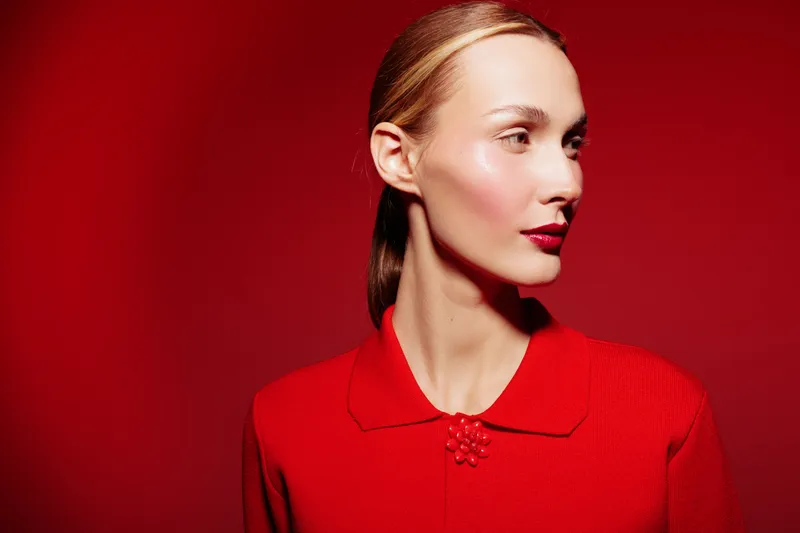 Portrait of a young woman with slicked hair wearing a red sweater against a red background. The image highlights fashion, beauty, and vibrant color contrast in studio lighting.