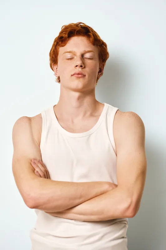 Young man with red hair and pale skin standing with crossed arms wearing white tank top. Closed eyes and relaxed expression on light background for portrait and confidence.