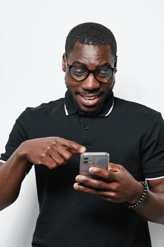 Black man with dark skin wearing glasses and black polo shirt smiling while using smartphone for texting or browsing on white background.