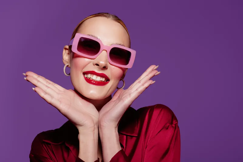 Smiling woman with pink sunglasses and red lipstick poses with hands near face on purple background. Concept of fashion, beauty, glamour, and confident female model portrait.