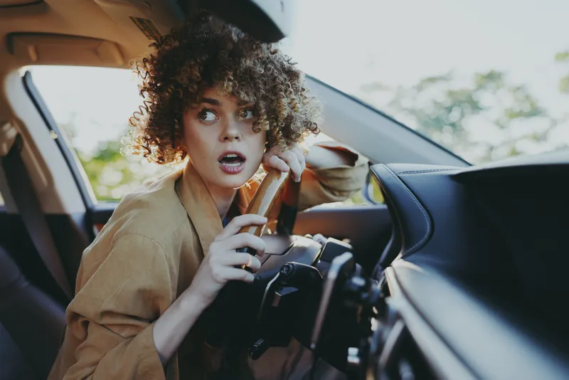 Young woman with curly hair sits in a car, surprised by something outside, casual daylight interior shot that captures a spontaneous, candid moment through the windshield.