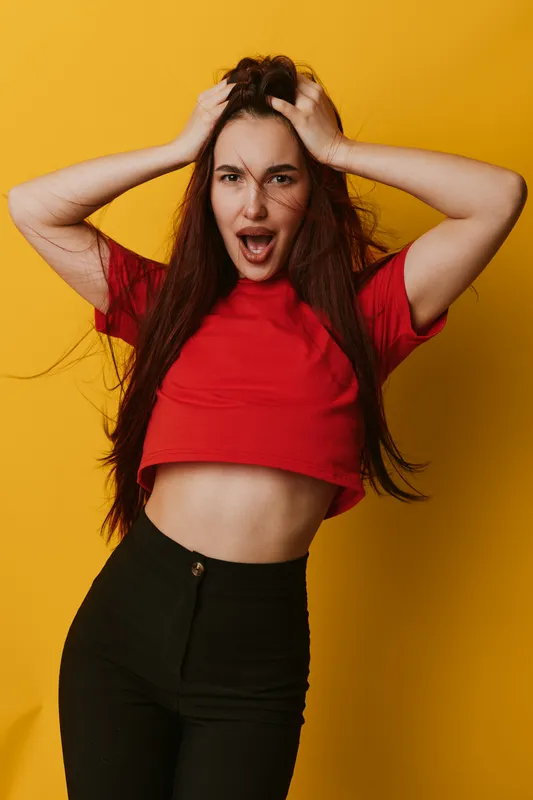 Young woman with long hair wearing red crop top and black pants posing with hands in hair and expressive face on yellow background. Casual style and emotional portrait in studio.