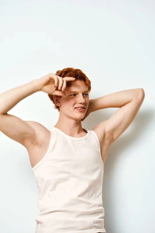 Young man with red hair and light skin wearing sleeveless white shirt posing with hands behind head on white background. Casual style and confident facial expression in studio light.