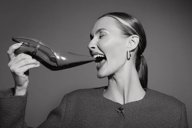 A smiling white woman with light skin holds a high heel shoe close to her mouth, playful expression in a black and white studio portrait. Concept of fashion and fun.