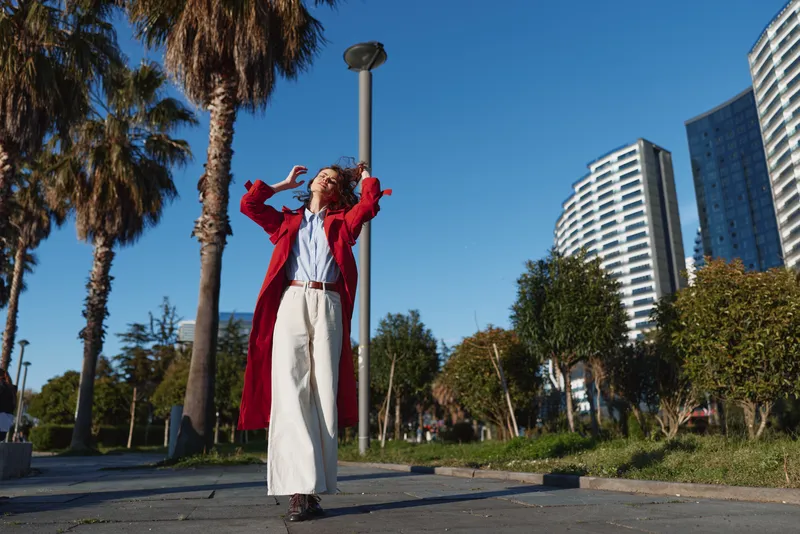 Confident woman in a red coat and white dress posing outdoors in an urban park with palm trees and modern buildings, showcasing bold fashion and sunny street style.