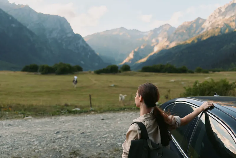 Young woman with ponytail leaning on car door looking at mountains in sunny nature landscape. Concept of travel, adventure, lifestyle, and outdoor exploration.