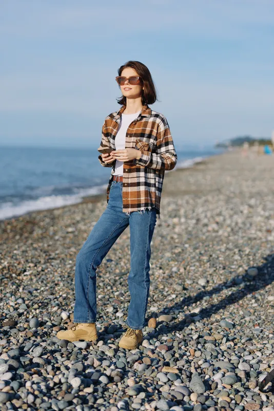 Woman standing on a pebble beach in casual attire, plaid shirt, denim jeans and boots, holding a cup and enjoying a sunny day by the sea with a clear sky above