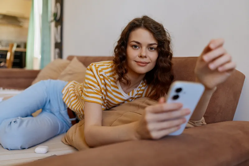 A relaxed woman lounges on a sofa at home, scrolling on a smartphone. Casual, cozy setting conveys comfortable, everyday life and personal downtime in a bright living room with warm light and soft