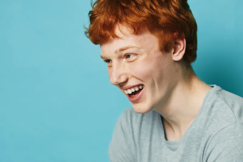 Young man with red hair smiling and looking away on blue background. Casual portrait of a happy male with freckles and comfortable clothing in studio lighting.