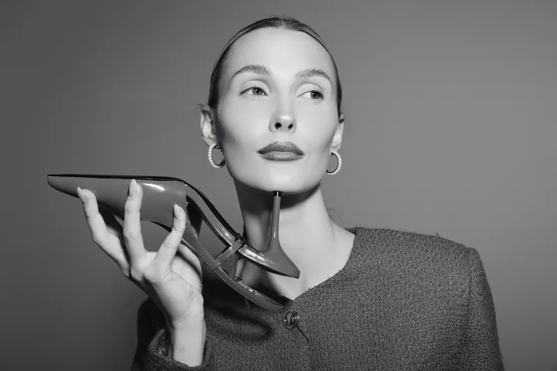 Portrait of a fair skinned white woman in jacket holding high heel shoe near face with confident expression. Fashion concept with minimalistic studio background.
