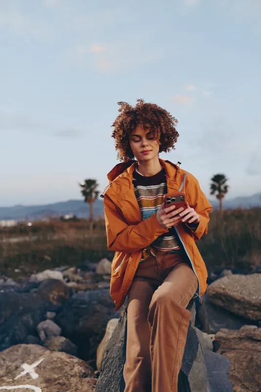 Woman lifestyle outdoors, curly hair, orange jacket, holding coffee cup, standing on rocks at sunrise, relaxed expression, soft sunlight, dry landscape, palm trees, natural colors