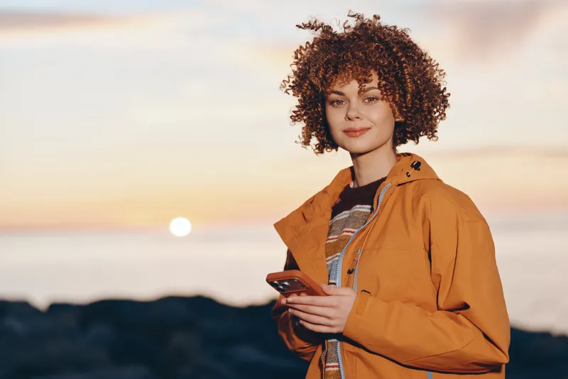Woman with curly hair wearing orange jacket holding a book outdoors at sunset, lifestyle portrait, soft ocean background, warm natural light, confident calm mood, by the sea