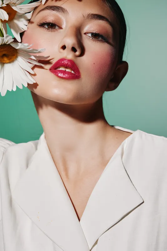 Close-up of a young woman with glossy red lipstick and vibrant blush holding large white flowers near her face on a vivid mint green background. Fashion beauty concept.