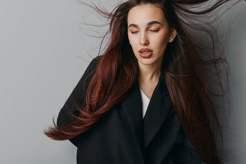 Woman with long brown hair and closed eyes wearing black coat on gray background. Portrait of young female with natural makeup and flowing hair in studio for fashion or beauty concept.