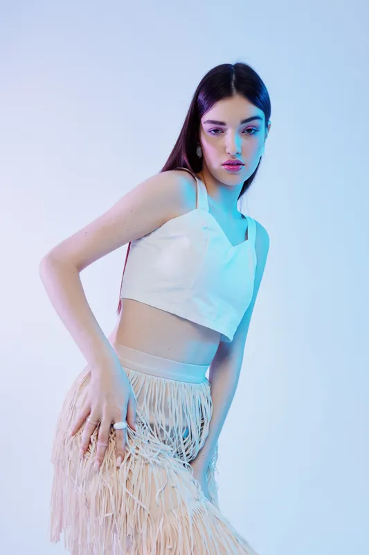 Young woman beauty fashion model posing in white crop top and fringed skirt against light background with soft blue and pink lighting. Concept of style and elegance.