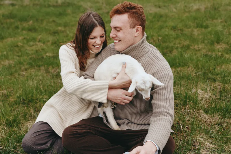 A joyful couple sit on grass, cuddling a white goat kid in a sunlit field. A warm, candid scene highlighting lifestyle, outdoor life, connection with animals and shared affection.