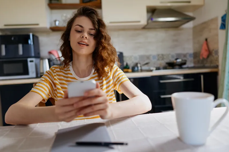 Young woman in a kitchen sits at a table using a smartphone, casual home lifestyle moment, bright light, relaxed mood, focused on device