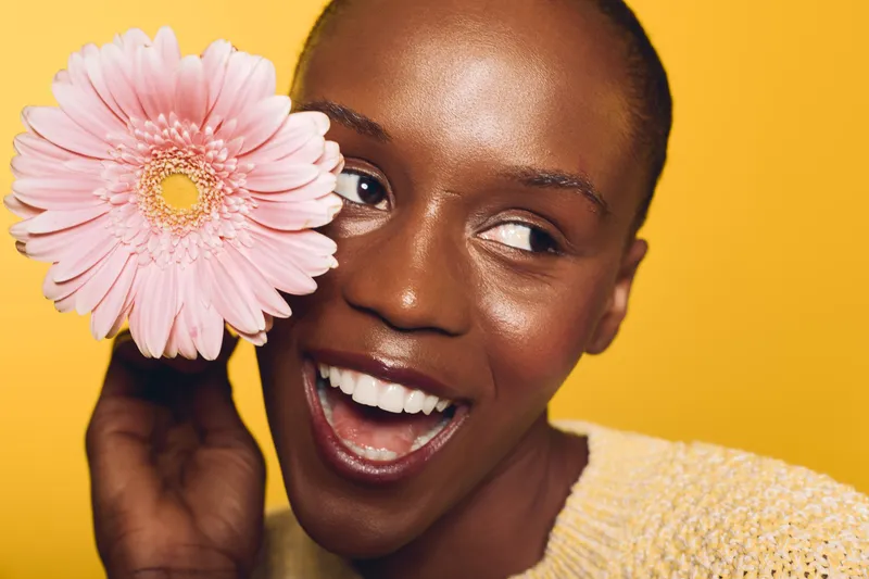 Close-up of happy African woman with dark skin holding pink flower near face against yellow background. Concept of beauty, skincare, natural smile, and positive mood in studio lighting.