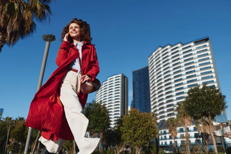 Young woman in a red coat walks confidently in a sunny urban park with modern high-rise buildings in the background, enjoying a bright and cheerful day outdoors.