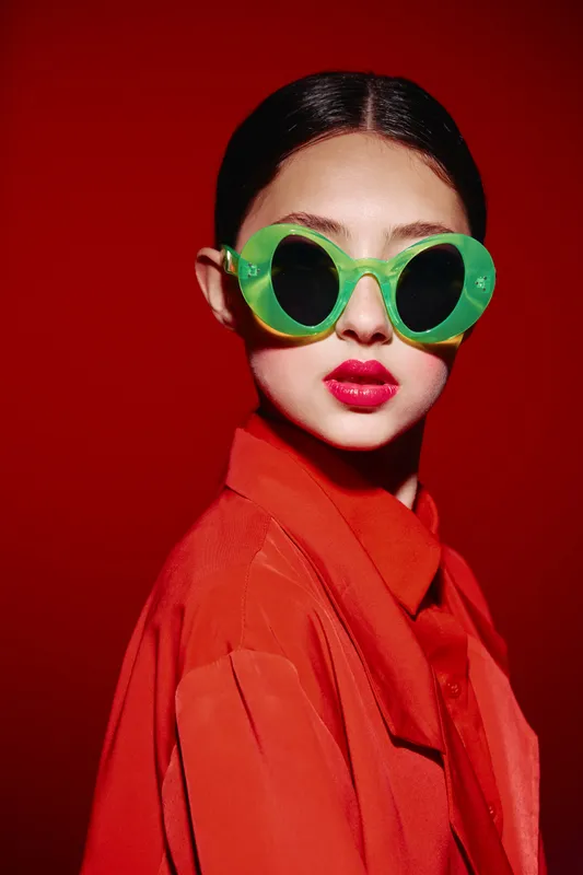 Fashion model wearing oversized green sunglasses and bright red lipstick dressed in orange coat on dark red background. Studio shot with strong contrast and creative mood.