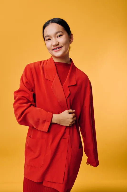 Young woman wearing oversized red jacket and matching dress standing against yellow background smiling with hand resting on jacket in casual fashion style portrait with confident expression