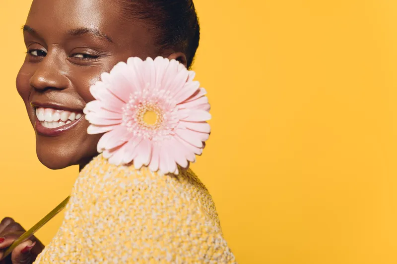Smiling black woman with dark skin holds pink flower near face against yellow background. Concept of natural beauty, happiness, skincare, and floral wellness with bright warm lighting.