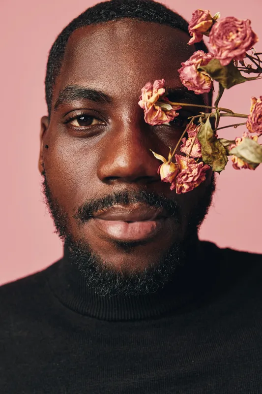 African man with dark skin and beard posing with dried pink flowers near face in creative conceptual visual style on pink background. Portrait with artistic expression and fashion.