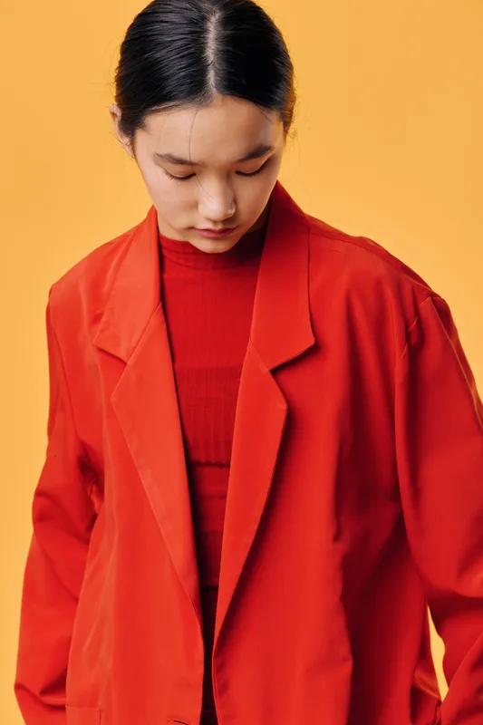 Young woman with black hair wearing red coat and turtleneck standing against yellow background looking down thoughtfully and quietly in fashion portrait style.
