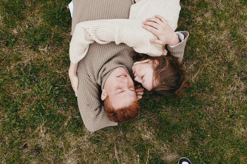 couple outdoors grass sweater cozy affection relaxed sunlight moment intimate, landscape park leisure lifestyle, closeup embrace smiling carefree breeze casual knitwear beige brown earthy tone nature