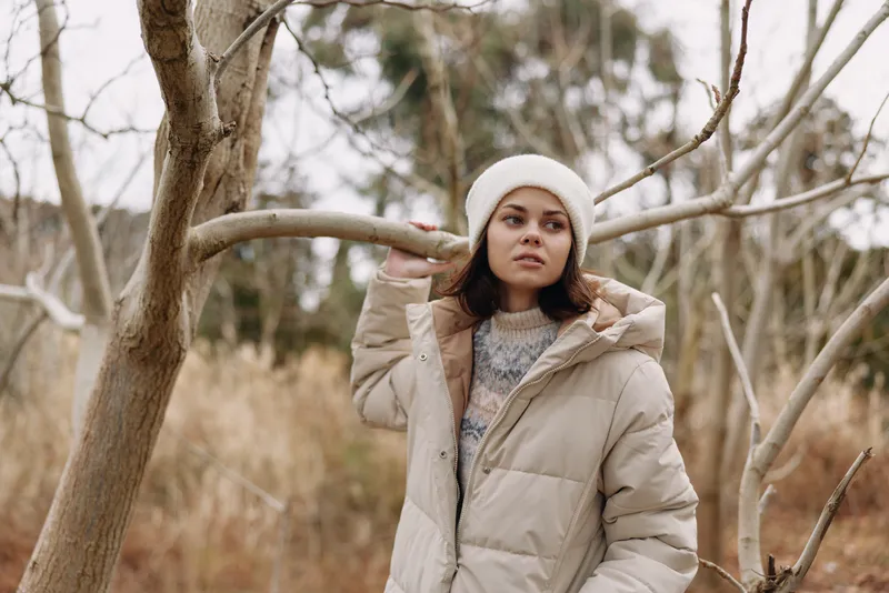 Woman in a warm coat and white beanie standing among bare trees in an outdoor winter portrait, serene mood, natural light, muted tones, and calm candid style.
