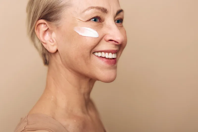 Smiling mature woman with short hair applies white cream on cheek, demonstrating skincare routine and self-care. Close-up portrait on beige background with natural light and healthy skin concept.