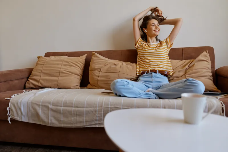 Young woman sits on a cozy sofa at home wearing a striped yellow T shirt and light jeans smiling as she runs her hands through her hair in a relaxed mood