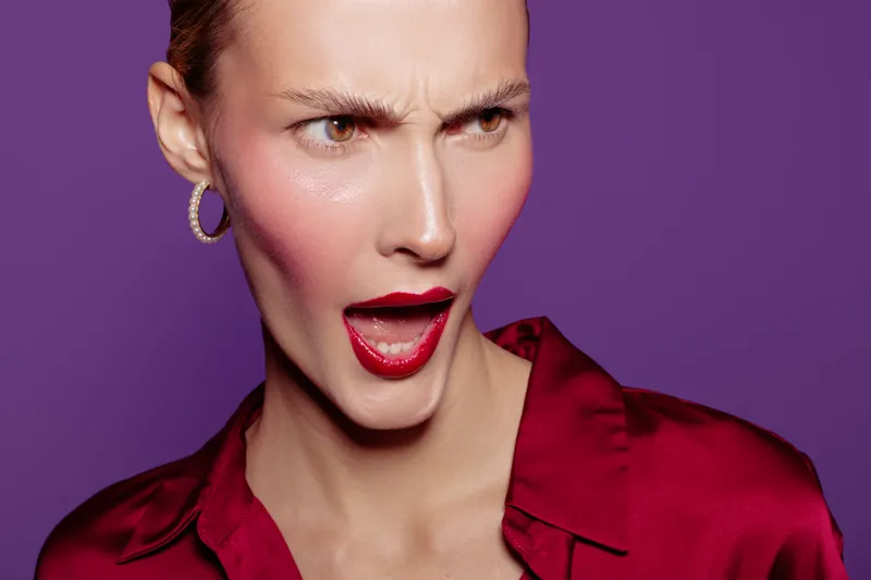 Closeup of surprised woman with red lipstick and hoop earrings wearing red satin blouse on purple background. Concept of emotion, expression, fashion, and beauty portrait.