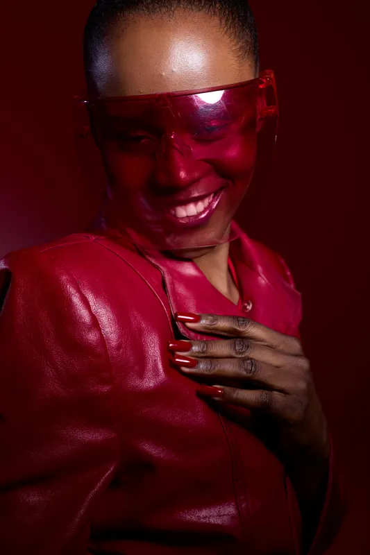 Smiling African woman with dark skin wearing red leather jacket and translucent red visor. Studio portrait with vibrant red lighting. Concept for fashion, style, confidence, and beauty.